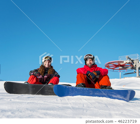 Guy and girl snowboarders are sitting on the snow at the top of the ski slope against the blue sky 46223719