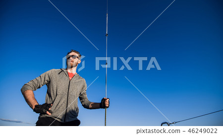 Young man on a catamaran standing and holding rope in front of a blue sky during cruising. 46249022