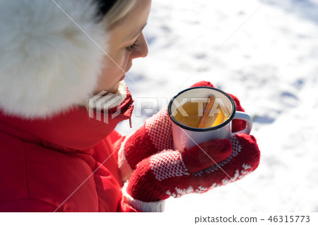 Hands in Knitted Mittens holding Steaming Cup of Hot Tea on Snowy Winter Morning Outdoors. Woman 46315773