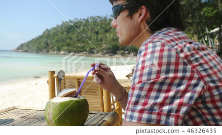 Handsome young man in sunglasses drink fresh coconut juice in a beach cafe with sea view on palm Handsome young man in sunglasses drink fresh coconut juice in a beach cafe with sea view on palm 46326435