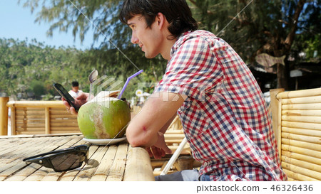 Handsome young businessman in a beach cafe with sea view on palm trees on background and uses mobile 46326436