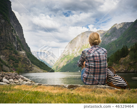 A woman sits on the shore of a picturesque fjord in Norway, admiring the beauty of Scandinavian A woman sits on the shore of a picturesque fjord in Norway, admiring the beauty of Scandinavian 46329464