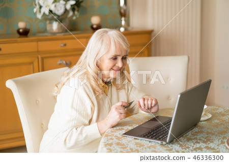 Smiling senior woman uses laptop and credit card while sitting at the table Smiling senior woman uses laptop and credit card while sitting at the table 46336300