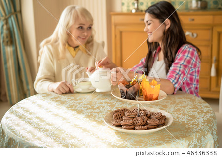 Senior mother drinks tea with her smiling daughter. Desserts stand on the table Senior mother drinks tea with her smiling daughter. Desserts stand on the table 46336338