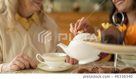 Hands of young woman with white china teapot pouring tea into cup of her senior mother Hands of young woman with white china teapot pouring tea into cup of her senior mother 46336340