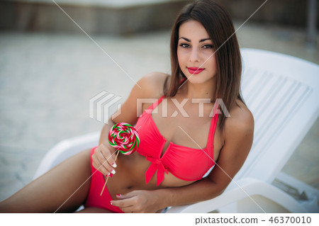A beautiful girl in a pink bathing suit sunbathing by the swimming pool. Girl hold candy. Sexy lady A beautiful girl in a pink bathing suit sunbathing by the swimming pool. Girl hold candy. Sexy lady 46370010