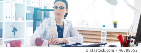 A beautiful young girl in a white robe is sitting at a table and holding a wooden figure of a man in 46399552