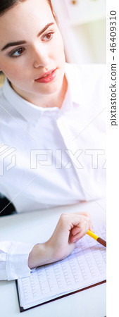 A young girl in the office holds a pen in her hand and works with documents and a computer. 46409310