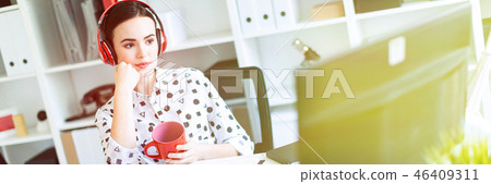 A young girl sits in headphones at a table in the office, holds a red cup in her hands and looks at 46409311
