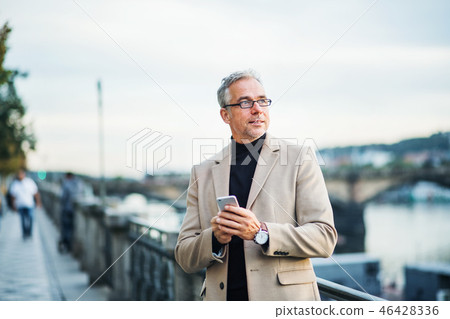 Mature businessman with smartphone standing by river in Prague city, using smartphone. 46428336