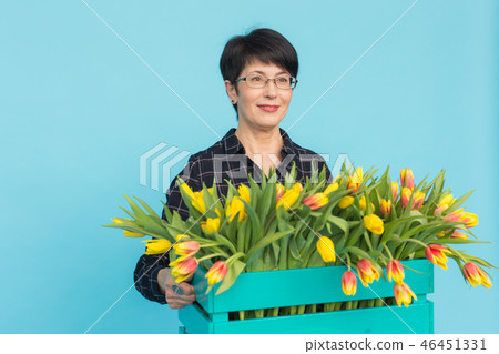 Happy middle-aged woman florist wearing glasses with box of tulips on blue background Happy middle-aged woman florist wearing glasses with box of tulips on blue background 46451331