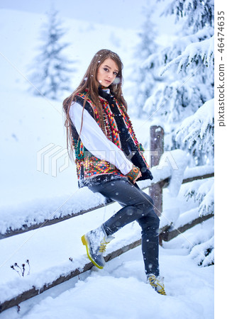 Fashionable girl in jeans, white embroidered blouse and multicolored woolen vest at wooden fence. 46474635