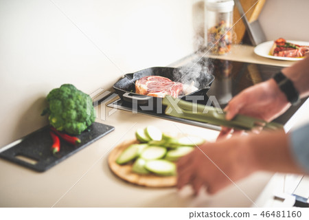 Piece of meat with steam frying on stovetop while man preparing vegetables 46481160