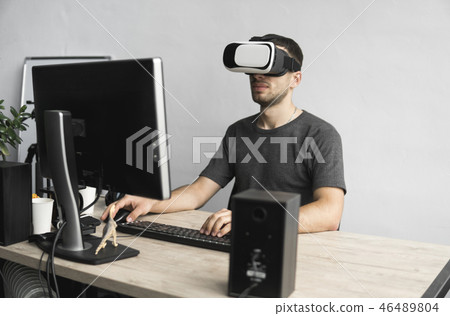 Young man wearing virtual reality goggles headset, vr box and sitting in the office against computer Young man wearing virtual reality goggles headset, vr box and sitting in the office against computer 46489804