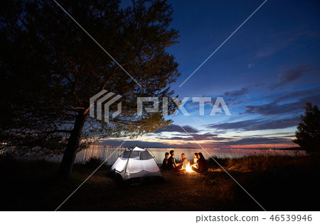 Night summer camping on shore. Group of young tourists around campfire near tent under evening sky 46539946