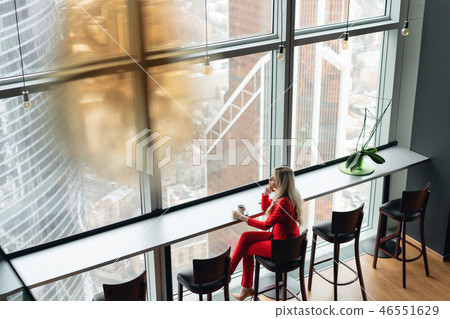 Woman in red coral business suit drinks coffee at a high table near the window. Young blonde 46551629