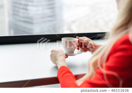 Cup of hot drink in hand close-up. Woman in red coral business suit drinks coffee at a high table 46551636