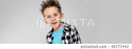 A handsome boy in a plaid shirt, blue shirt and jeans stands on a gray background. The boy folded 46573442