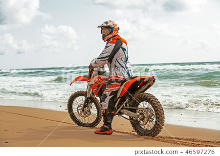 Motorcyclist in a protective suit sitting on motorbike in front of the sea Motorcyclist in a protective suit sitting on motorbike in front of the sea 46597276