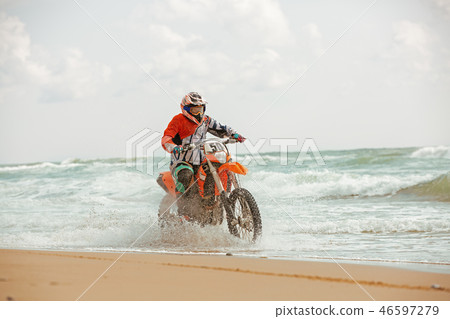 Motorcyclist in a protective suit rides a motorcycle on the sea, splashes fly from under the wheels. Motorcyclist in a protective suit rides a motorcycle on the sea, splashes fly from under the wheels. 46597279