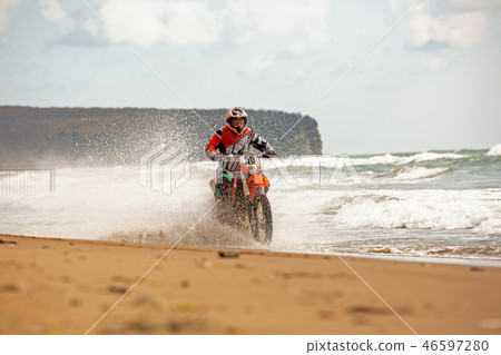Motorcyclist in a protective suit rides a motorcycle on the sea, splashes fly from under the wheels. Motorcyclist in a protective suit rides a motorcycle on the sea, splashes fly from under the wheels. 46597280