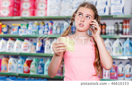 serious girl standing in store choosing products while talking by phone and looking at shopping list 46610153