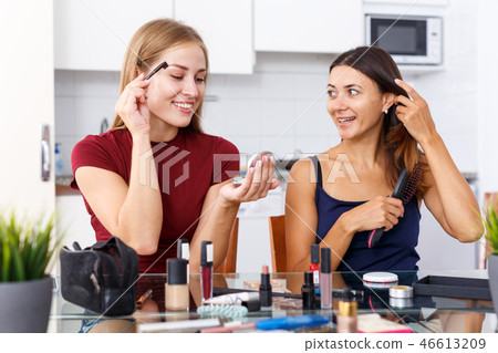 Two positive young girl friends doing make up at table with cosmetics Two positive young girl friends doing make up at table with cosmetics 46613209