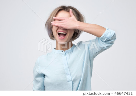 Happy young woman in blue shirt covering her eyes over gray background Happy young woman in blue shirt covering her eyes over gray background 46647431