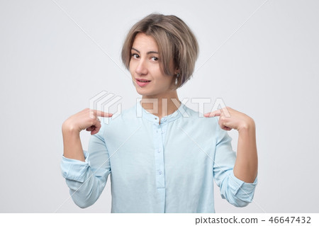 Pretty young woman in blue shirt pointing at herself in confusion Pretty young woman in blue shirt pointing at herself in confusion 46647432
