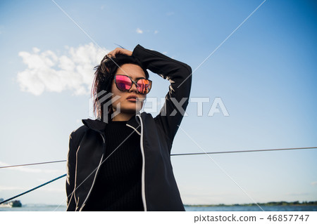 Young smiling dark-haired woman wearing sunglasses sitting on a board of boat in sunny autumn day Young smiling dark-haired woman wearing sunglasses sitting on a board of boat in sunny autumn day 46857747