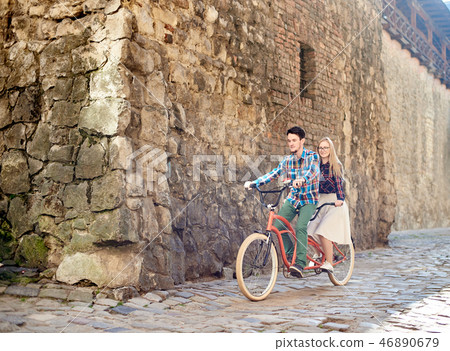Young tourist couple, handsome man and pretty blond woman riding tandem bicycle along city street. 46890679
