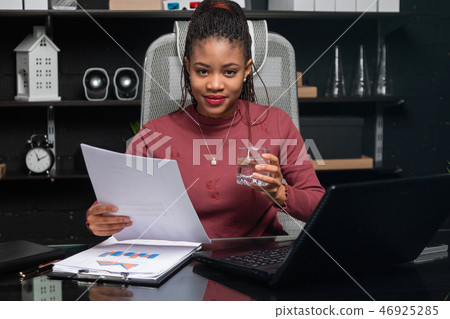 Beautiful young African-American businesswoman holding glass of water and working with documents at Beautiful young African-American businesswoman holding glass of water and working with documents at 46925285
