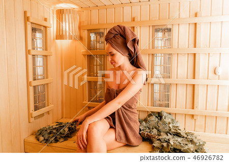Young woman relaxing in a sauna dressed in a towel. Interior of new Finnish sauna, infrared panels Young woman relaxing in a sauna dressed in a towel. Interior of new Finnish sauna, infrared panels 46926782