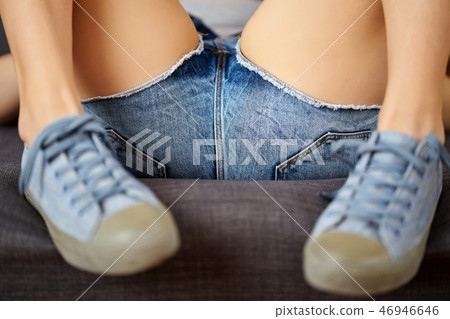 View of sexy female legs in jeans shorts and blue sneakers on a gray couch. View of sexy female legs in jeans shorts and blue sneakers on a gray couch. 46946646