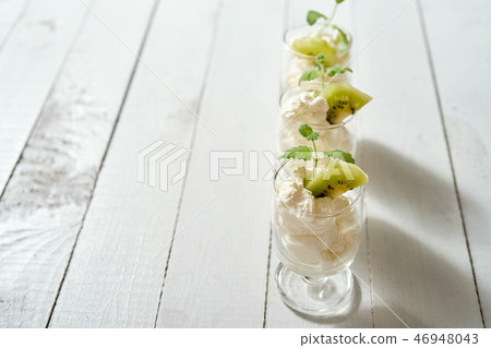 Glass of dessert with whipped cream with a fresh mint leaf and kiwi fruit on white table. 46948043