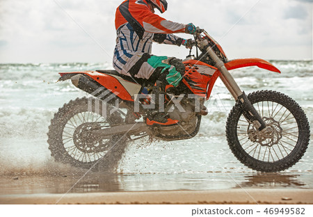 Motorcyclist in a protective suit rides a motorcycle on the sea, splashes fly from under the wheels. 46949582