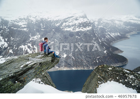 Trolltunga cliff under snow in Norway. Scenic Landscape. Man traveller sitting on edge of rock and Trolltunga cliff under snow in Norway. Scenic Landscape. Man traveller sitting on edge of rock and 46957678