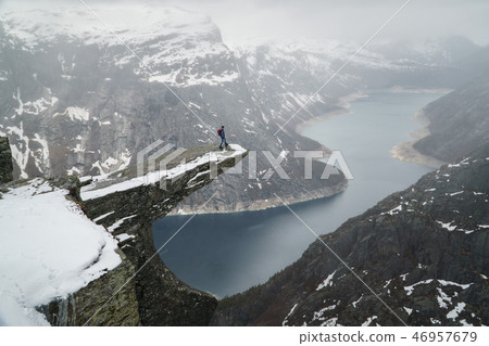 Trolltunga cliff under snow in Norway. Scenic Landscape. Man traveller standing on edge of rock and Trolltunga cliff under snow in Norway. Scenic Landscape. Man traveller standing on edge of rock and 46957679