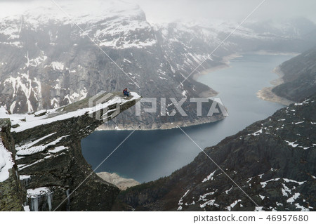 Trolltunga cliff under snow in Norway. Scenic Landscape. Man traveller sitting on edge of rock in Trolltunga cliff under snow in Norway. Scenic Landscape. Man traveller sitting on edge of rock in 46957680