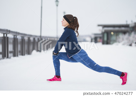 Young female runner doing flexibility exercise for legs before run at snow winter promenade Young female runner doing flexibility exercise for legs before run at snow winter promenade 47024277