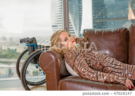 A beautiful young blonde girl in a fashionable dress with a disability, posing on a leather sofa 47028147