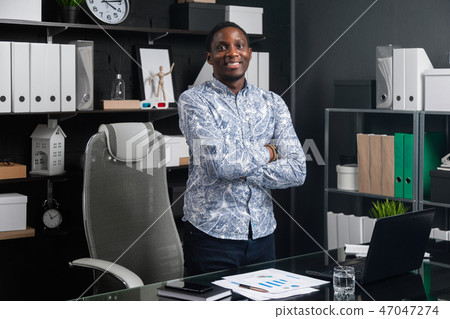 portrait of young black businessman standing near table in office with his hands folded on his chest portrait of young black businessman standing near table in office with his hands folded on his chest 47047274