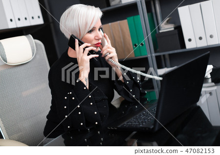 Busy woman talking on mobile and landline sitting at table in office 47082153
