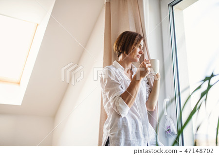 A young woman with night shirt standing by the window in the morning, holding cup of coffee. A young woman with night shirt standing by the window in the morning, holding cup of coffee. 47148702