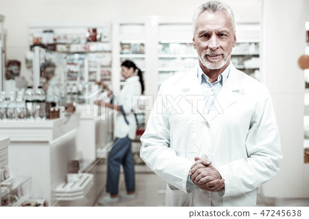 Bearded pharmacist wearing white coat looking pleasant meeting his clients Bearded pharmacist wearing white coat looking pleasant meeting his clients 47245638