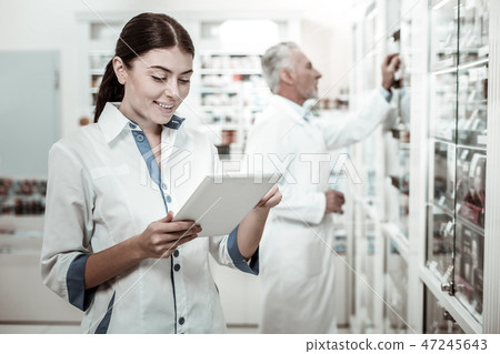 Beautiful dark-haired pharmacist holding tablet while looking at medicine range Beautiful dark-haired pharmacist holding tablet while looking at medicine range 47245643