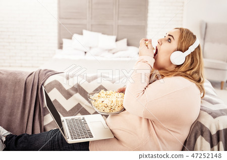 Overweight young woman with headphones on unpleasantly eating Overweight young woman with headphones on unpleasantly eating 47252148