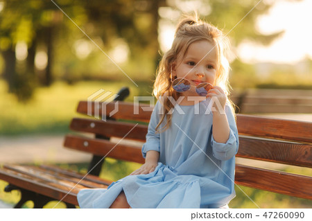 Little girl model in blue dress and sun glasses sits on a bench in the park 47260090