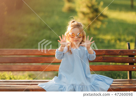 Little girl model in blue dress and sun glasses sits on a bench in the park Little girl model in blue dress and sun glasses sits on a bench in the park 47260098