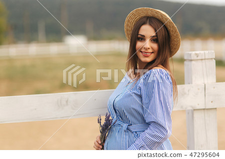 Beautiful pregnant woman in blue dress walking near the farm. Knitted bag and bouquet of lavender Beautiful pregnant woman in blue dress walking near the farm. Knitted bag and bouquet of lavender 47295604